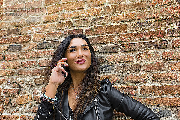 Porträt einer jungen Frau in schwarzer Lederjacke  mit Smartphone  im Hintergrund Backsteinmauer