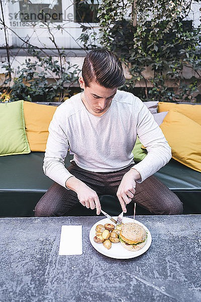 Junger Mann sitzt in einem Restaurant auf der Couch und isst einen veganen Burger zu Mittag