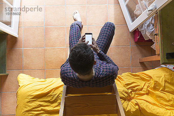 Mann mit Smartphone  der im Schlafzimmer auf einer Holzleiter sitzt