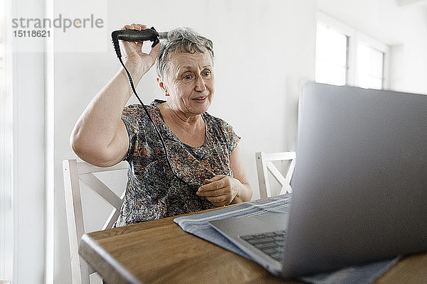 Ältere Frau sitzt zu Hause am Tisch mit Laptop und macht sich die Haare