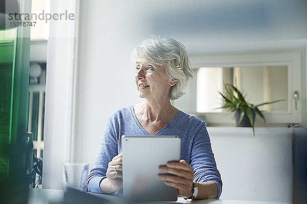 Lächelnde reife Frau mit digitalem Tablett  die aus dem Fenster schaut