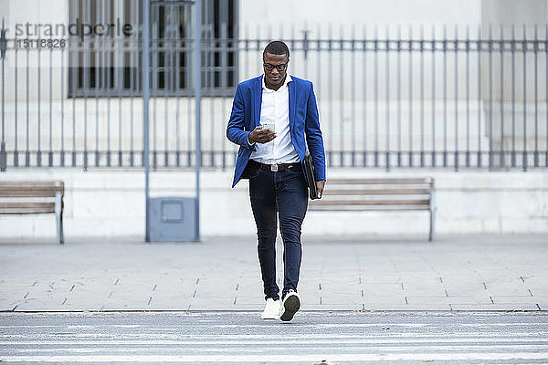 Junger Geschäftsmann in blauer Anzugjacke überquert die Straße und benutzt ein Smartphone