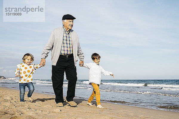 Großvater  der mit seinen Enkelkindern Hand in Hand am Strand spazieren geht