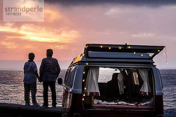 Älteres Ehepaar reist in einem Oldtimer-Van und betrachtet den Sonnenuntergang am Meer