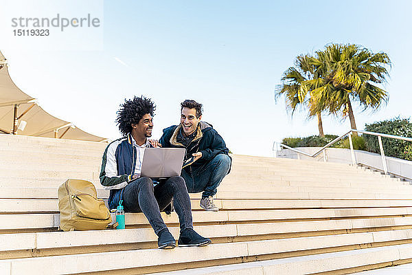 Zwei glückliche Geschäftsleute sitzen auf einer Treppe und arbeiten an einem Laptop