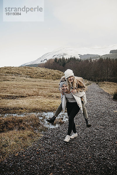 Grossbritannien  Schottland  glückliche junge Frau trägt Freundin huckepack in ländlicher Landschaft