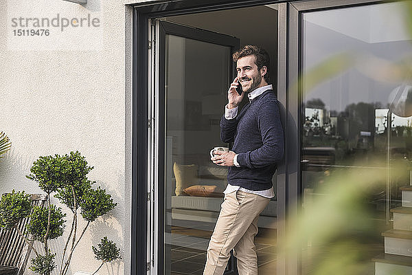 Junger Mann lehnt in Tür seines Hauses  hält Tasse Kaffee in der Hand  benutzt Smartphone
