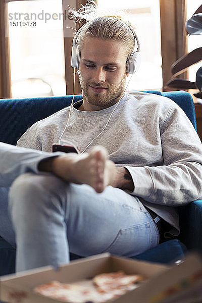Junger Mann sitzt auf Couch mit Handy und Kopfhörern