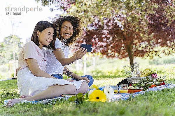 Zwei glückliche Frauen mit Handy und Kopfhörern im Park