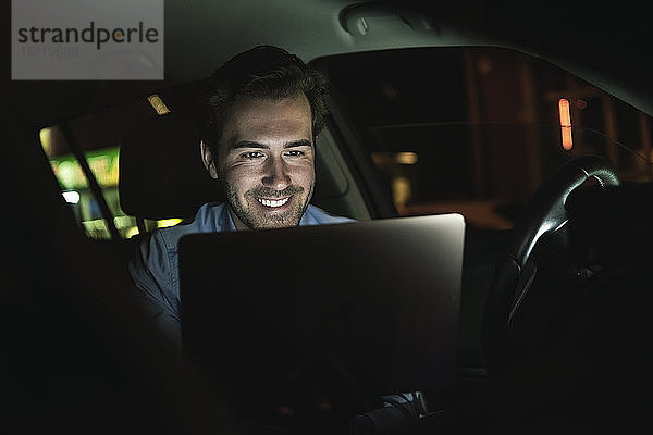 Lächelnder junger Mann mit Laptop nachts im Auto