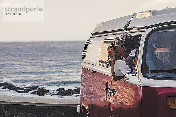 Ältere Frau sitzt in einem Oldtimer-Van und fotografiert das Meer