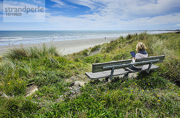 Frau sitzt auf einer Bank mit Tablette am Strand von Foxton  Kapiti Coast  Nordinsel  Neuseeland