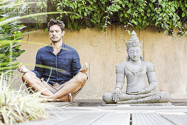 Mann sitzt im Schneidersitz neben der Buddhastatue in einem Zen-Garten und meditiert