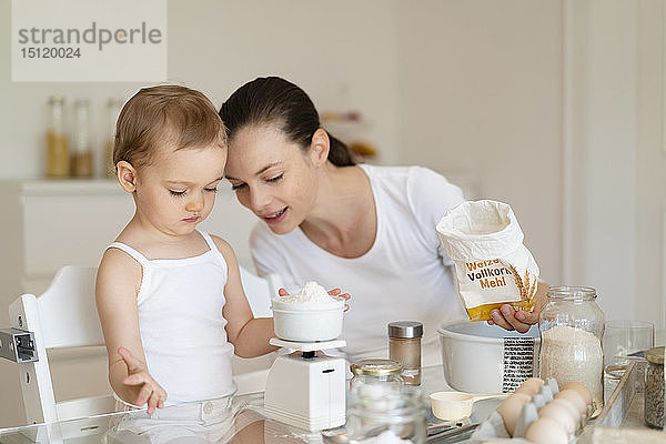 Mutter und kleine Tochter backen gemeinsam zu Hause in der Küche einen Kuchen und wiegen Mehl