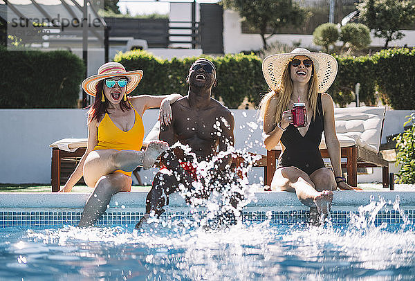 Zwei glückliche Frauen und ein Mann sitzen am Pool und spritzen Wasser
