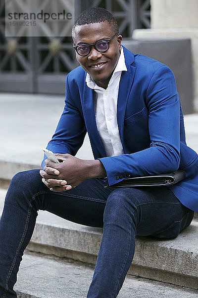 Junger Geschäftsmann in blauer Anzugjacke  sitzt auf einer Stufe und hält ein Smartphone