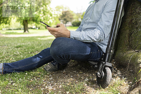 Mann sitzt auf einem E-Scooter in einem Park und lehnt sich an einen Baumstamm  während er ein Smartphone benutzt  Teilansicht