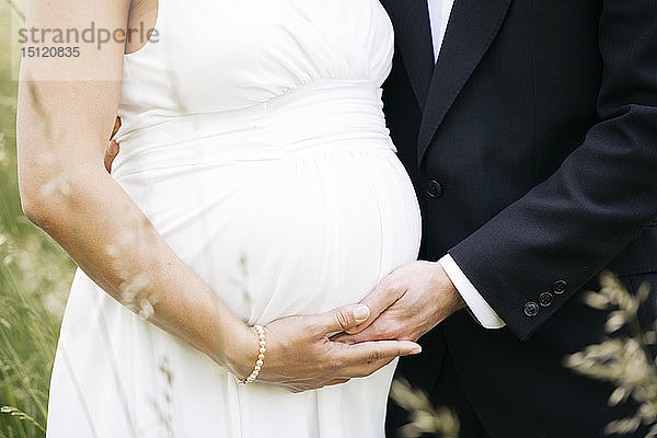 Schwangere Braut mit ihrem Ehemann hält Babybauch auf einer Wiese