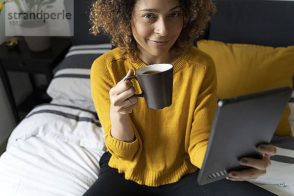Frau sitzt im Bett  macht eine Pause  trinkt Kaffee  tritt auf ein digitales Tablet