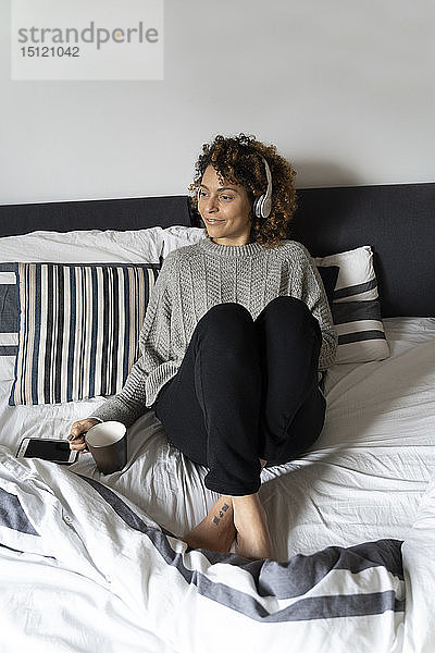 Frau sitzt auf dem Bett  trinkt Kaffee  hört Musik mit Kopfhörern und Smartphone