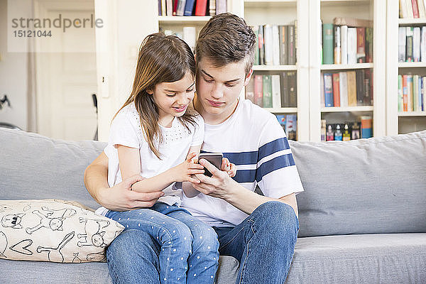Teenager  der mit seiner kleinen Schwester zu Hause auf der Couch sitzt und auf sein Handy schaut
