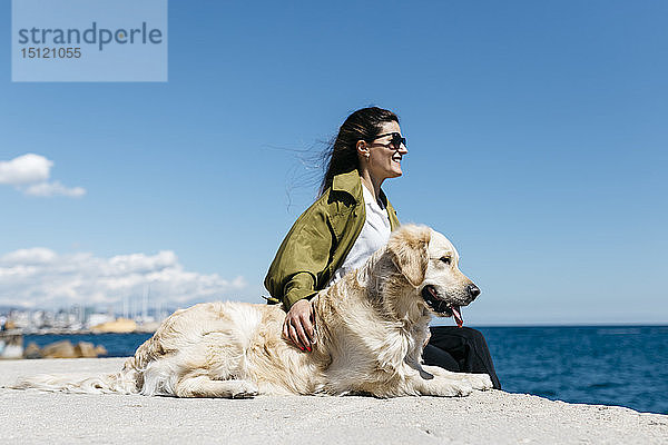 Glückliche Frau sitzt neben ihrem Labrador Retriever am Kai und schaut auf das Meer