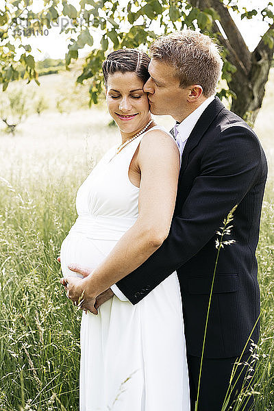 Schwangere Braut mit ihrem Ehemann hält Babybauch auf einer Wiese