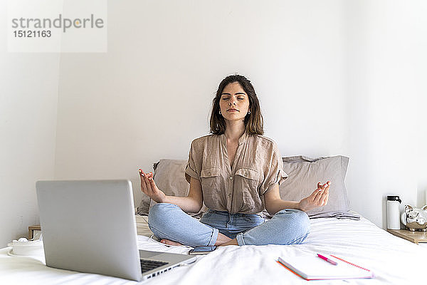 Junge Frau sitzt mit Laptop auf dem Bett und übt Yoga