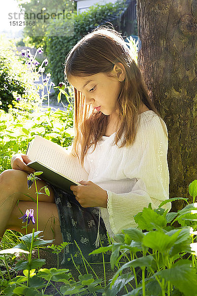 Kleines Mädchen lehnt am Baumstamm und liest ein Buch