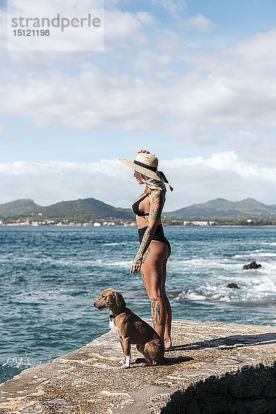Spanien  Mallorca  tätowierte Frau steht neben ihrem Hund und schaut auf das Meer
