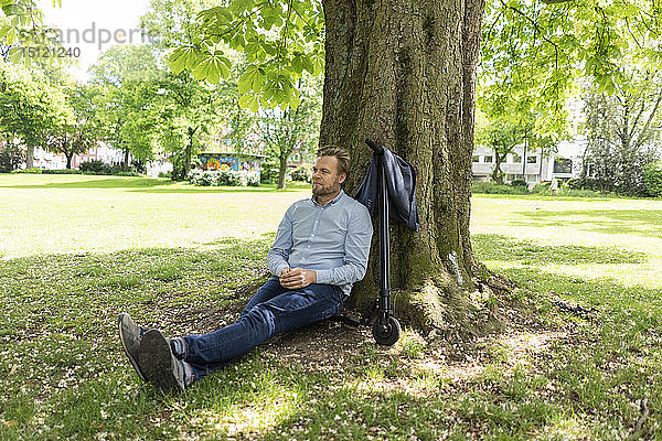 Geschäftsmann sitzt auf einem E-Scooter in einem Park und lehnt sich entspannt an einen Baumstamm