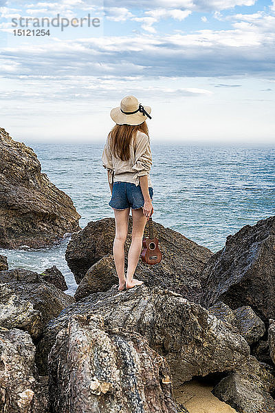 Rückenansicht einer jungen Frau mit Ukulele  die auf einem Felsen steht und auf das Meer schaut