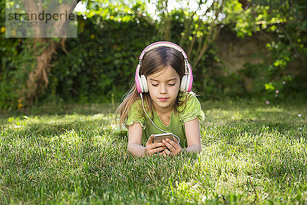 Porträt eines kleinen Mädchens  das auf einer Wiese liegt und mit Kopfhörern über ein Smartphone Musik hört