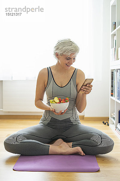 Frau  die zu Hause Yoga praktiziert  einen gesunden Fruchtsnack zu sich nimmt und ein Smartphone benutzt