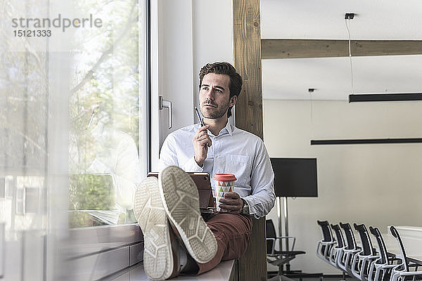Junger Geschäftsmann sitzt auf der Fensterbank  benutzt digitales Tablet  trinkt Kaffee