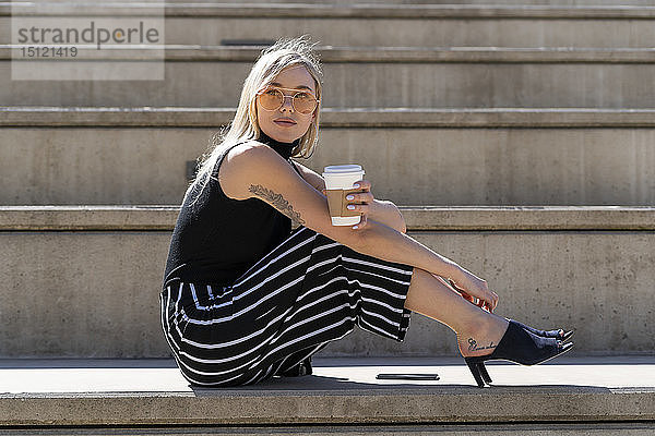 Porträt einer blonden jungen Frau mit Kaffee zum Sitzen auf einer Treppe im Freien