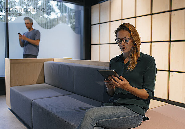 Junge Frau sitzt auf Couch mit Tablette mit Mann im Hintergrund