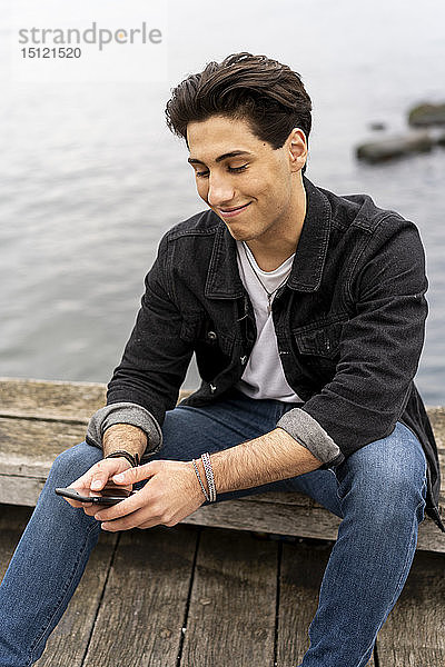 Dänemark  Kopenhagen  lächelnder junger Mann sitzt am Wasser und benutzt sein Handy