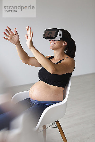 Junge schwangere Frau  die in einem Sessel sitzt und ihr Baby mit Hilfe einer Virtual-Reality-Brille beobachtet und ertastet