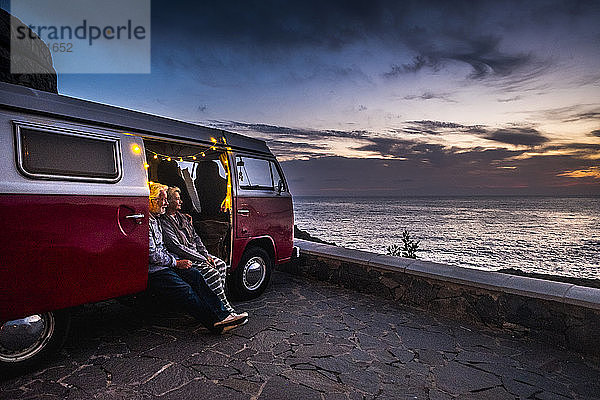 Älteres Ehepaar reist in einem Oldtimer-Van und betrachtet den Sonnenuntergang am Meer