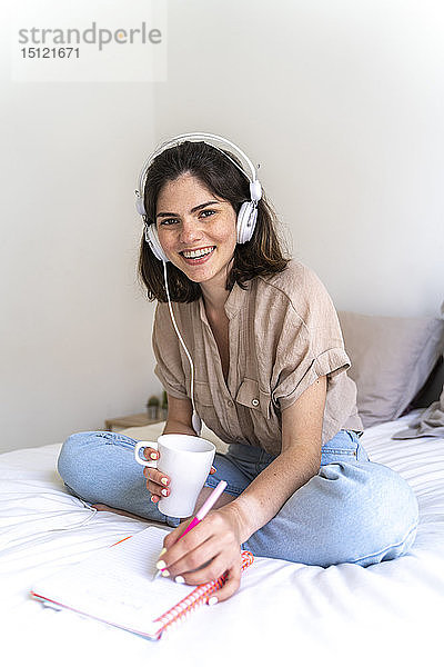 Porträt einer glücklichen jungen Frau  die mit Kopfhörern auf dem Bett sitzt und Notizen macht