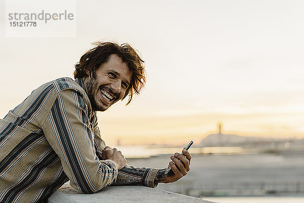 Porträt eines lachenden Mannes mit Smartphone bei Sonnenuntergang  Barcelona  Spanien