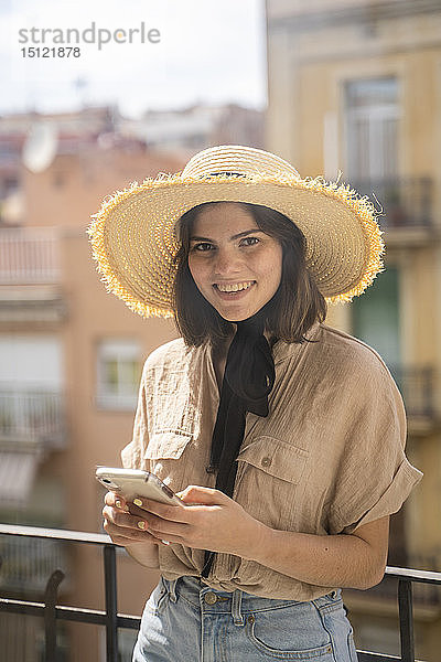Porträt einer lächelnden jungen Frau mit Strohhut  die auf einem Balkon in der Stadt steht