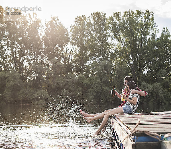 Glückliches junges Paar trinkt und planscht mit Wasser auf einem Steg an einem abgelegenen See