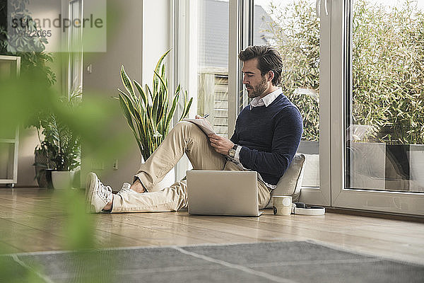 Junger Mann sitzt im Wohnzimmer  lehnt am Fenster  arbeitet mit dem Laptop  macht sich Notizen