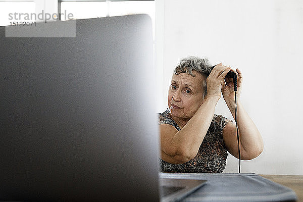 Ältere Frau sitzt zu Hause am Tisch mit Laptop und macht sich die Haare