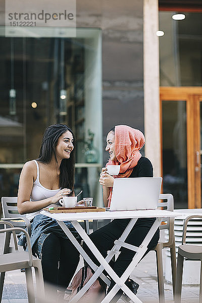 Zwei glückliche Freunde sitzen mit Laptop in einem Straßencafé zusammen
