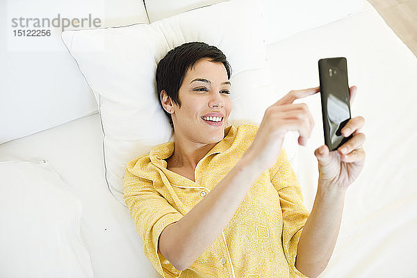 Lächelnde junge Frau benutzt Mobiltelefon im Bett