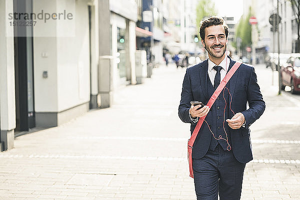 Lächelnder Geschäftsmann  der mit Handy und Kopfhörern durch die Stadt läuft