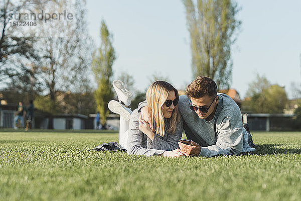 Junges Paar mit Sonnenbrille liegt auf einer Wiese und schaut auf sein Handy